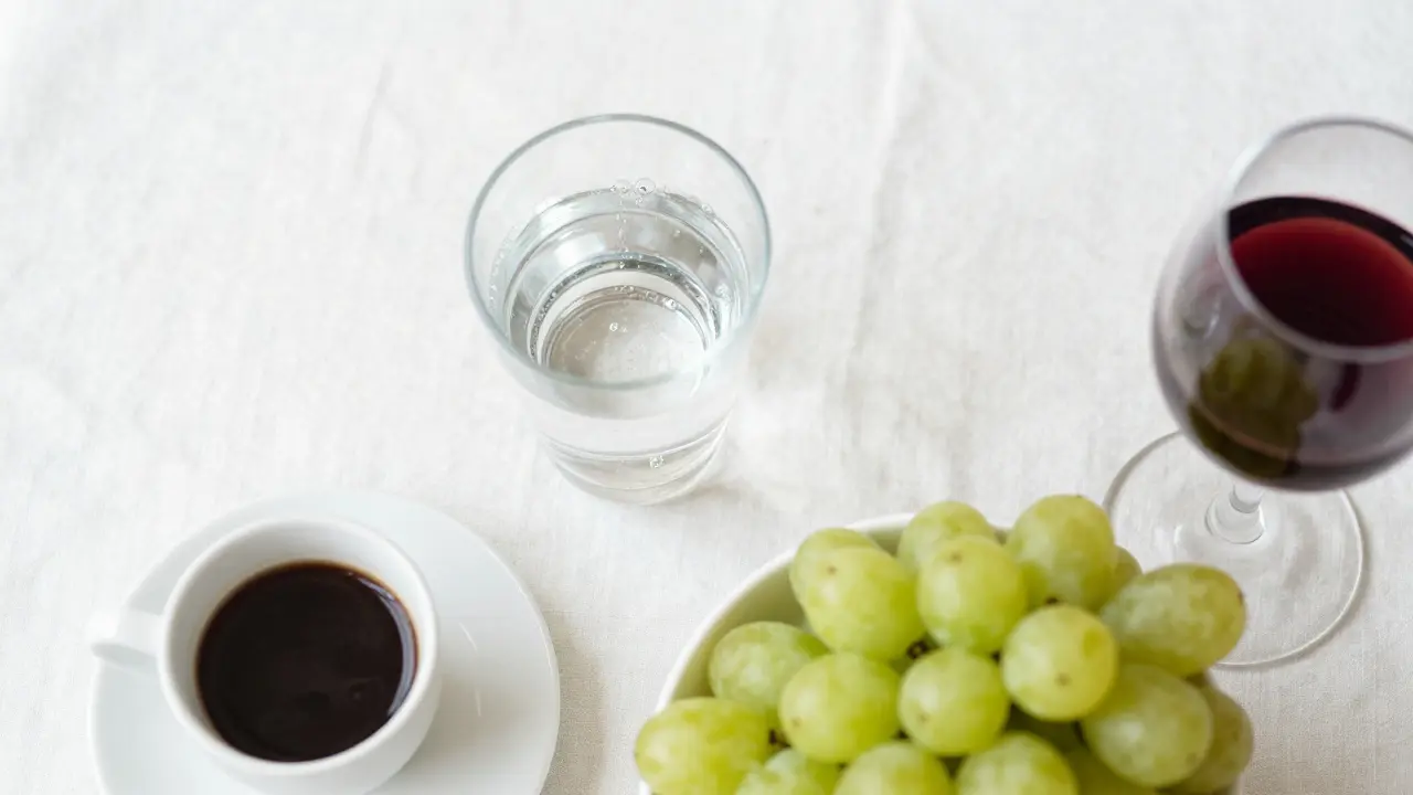 Stále voda a bílé hrozny vedle kávy a červeného vína na bílém ubrusu.